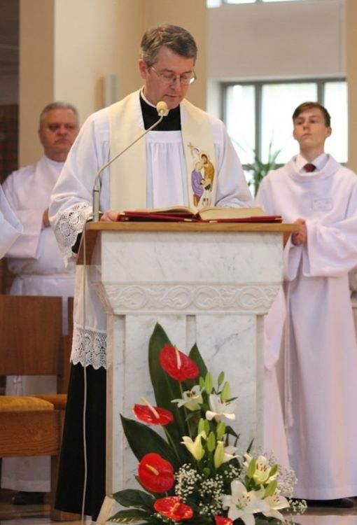 Ksiądz głoszący kazanie w kremowej stule liturgicznej z haftem Świętej Rodziny u góry i symbolami eucharystycznymi na dole, wykończonej złotymi frędzlami.