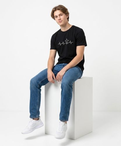 Młody mężczyzna w czarnej koszulce chrześcijańskiej z nadrukiem EKG i krzyża, niebieskich jeansach i białych trampkach, siedzący na jasnym postumencie